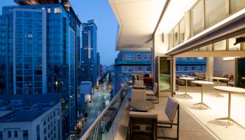 A modern cityscape viewed from a balcony at dusk, with tables and chairs, overlooking a street lined with tall buildings and illuminated windows.