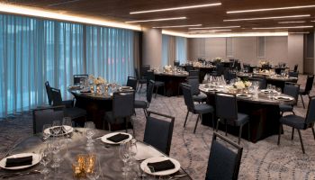 A banquet hall set up with round tables, black chairs, and elegant table settings, including napkins and glasses, under modern lighting fixtures.