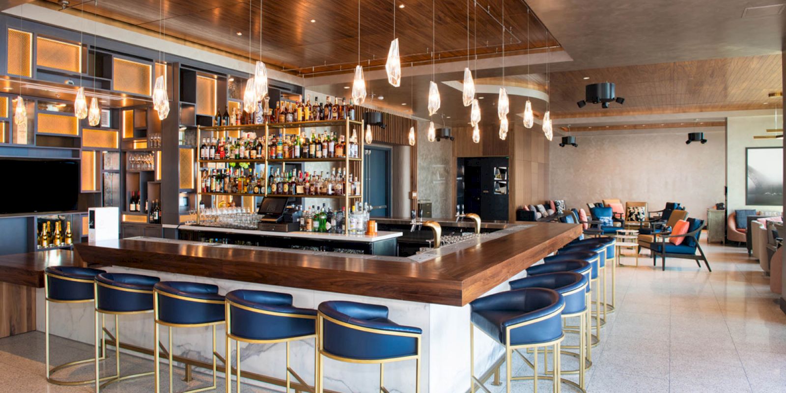 A modern bar with a wooden counter, numerous bottles on shelves, blue chairs, hanging lights, and an adjacent lounge area with sofas.