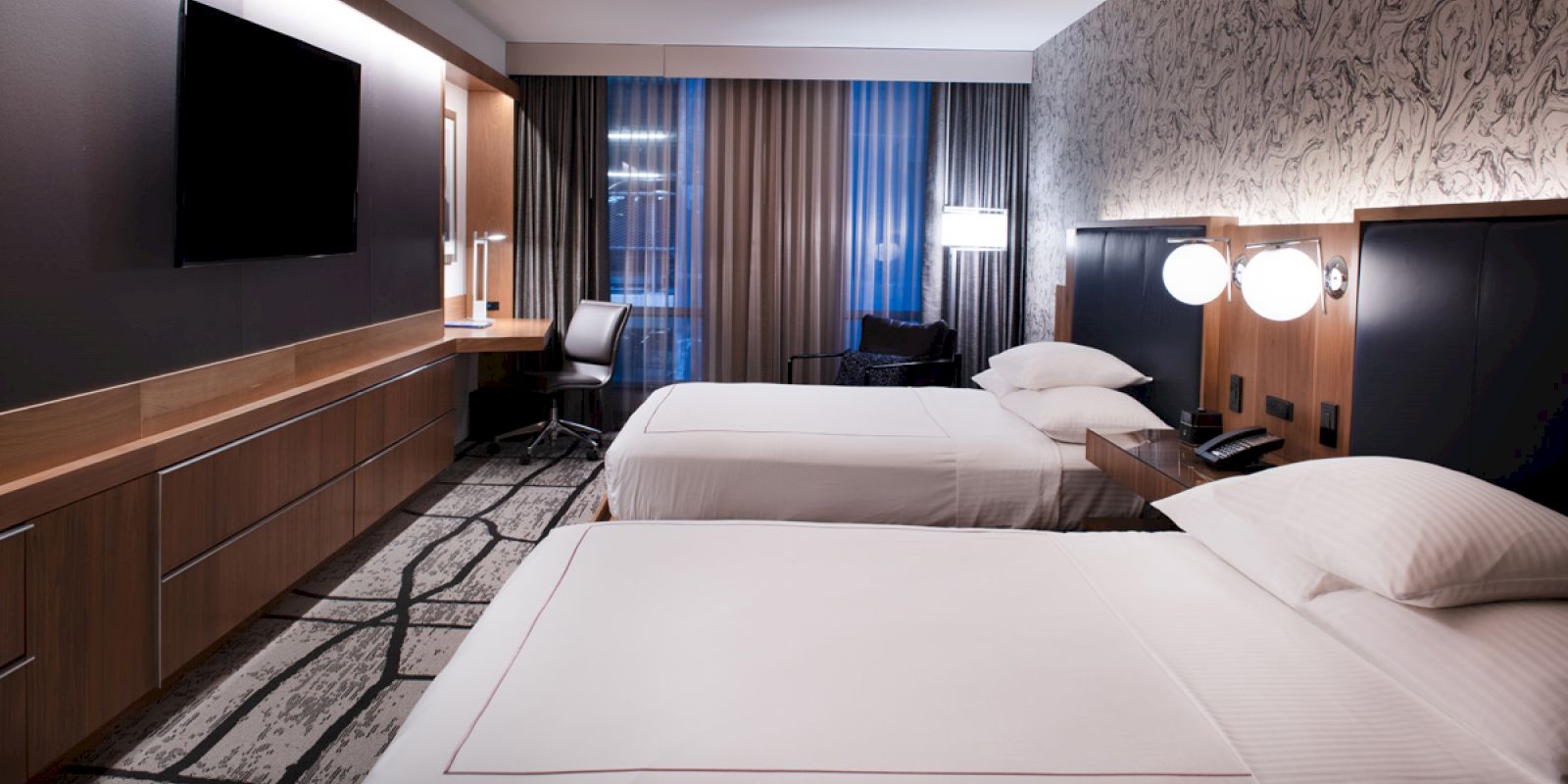 The image shows a modern hotel room with two neatly made beds, a mounted flat-screen TV, a desk, and contemporary decor within a well-lit space.