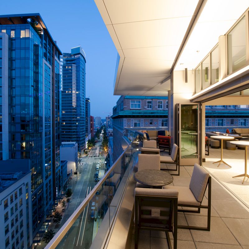 A modern cityscape is viewed from a high-rise balcony with tables and chairs, overlooking buildings and a street below, at dusk.