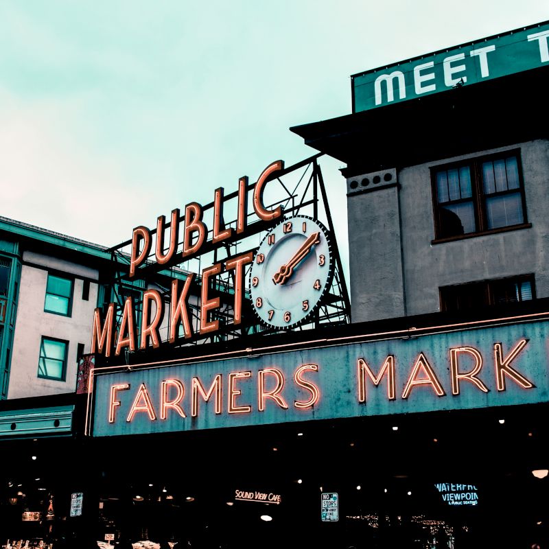 Image of a "Public Market" and "Farmers Market" sign with neon lights, and buildings in the background, including one labeled "PARKER HOTEL.".