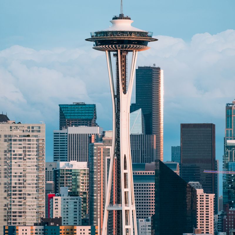This image shows the Space Needle, a prominent tower in Seattle, with various skyscrapers and buildings in the background under a partly cloudy sky.