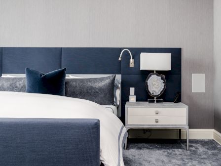 A contemporary bedroom with a large bed, navy headboard, and white bedding, a bedside table with a lamp and decorative item on it, a light gray rug.