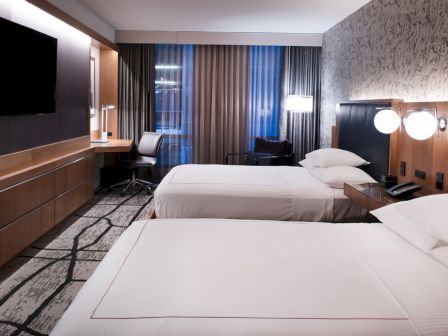 A modern hotel room with two double beds, a desk, a chair, a wall-mounted TV, and stylish decor, including a patterned accent wall and lamps.