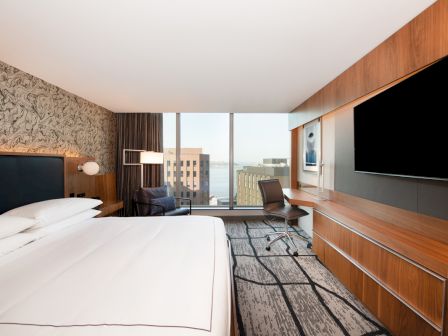 The image shows a modern hotel room with a large bed, a desk, a flat-screen TV, and a window with a city view in the background.