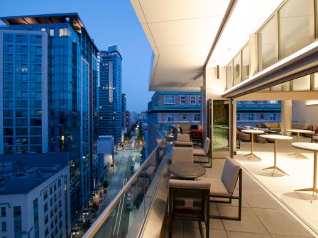 The image shows a modern rooftop terrace with seating, overlooking a cityscape with tall buildings at dusk. The skyline is illuminated by city lights.
