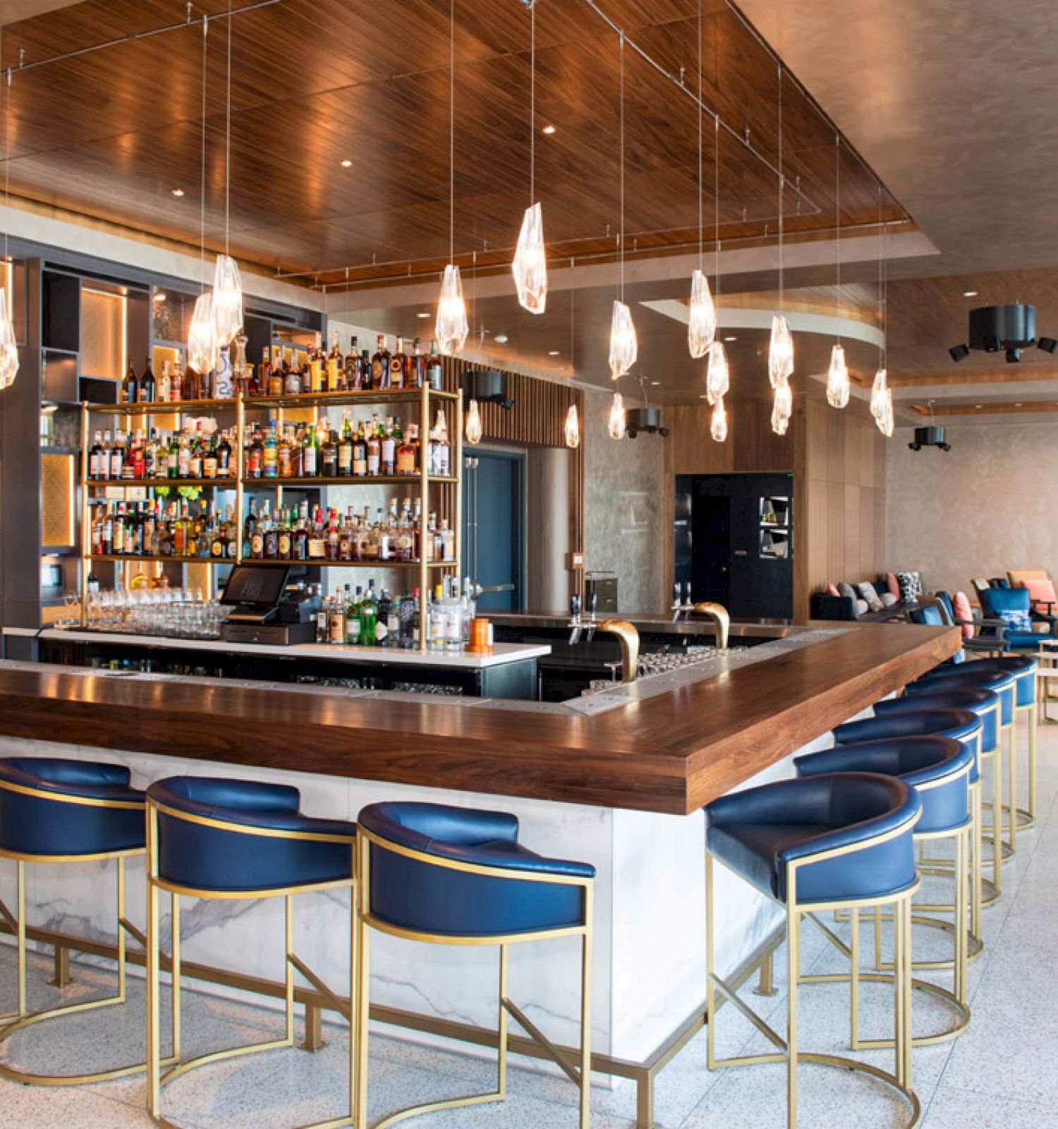 The image shows a modern bar interior with a wooden counter, stylish bar stools, a well-stocked bar, and contemporary seating in the background.