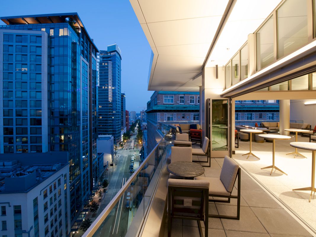 A modern balcony with seating and tables overlooks a cityscape of tall buildings at dusk, with lights illuminating the scene.