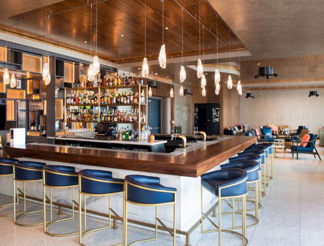 A modern bar with wooden accents, hanging lights, a variety of bottles on display, and chairs around the counter; a lounge area is in the background.