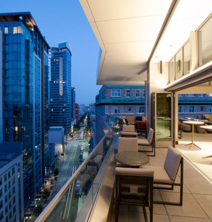 A modern balcony with seating overlooks a city street lined with tall buildings at dusk. The indoor area has tables and minimalistic decor.