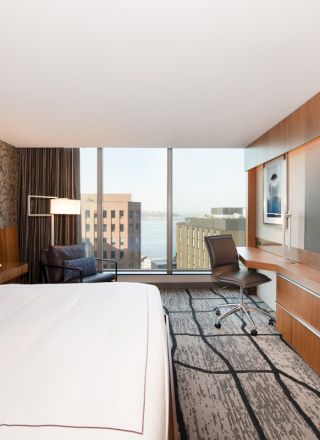 A modern hotel room featuring a large bed, wall-mounted TV, desk, chair, and a view of buildings and water through large windows ends the sentence.
