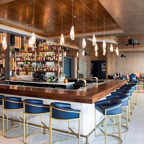 A stylish bar with a wooden counter, blue barstools, hanging lights, and a well-stocked liquor shelf, located in a modern lounge space.