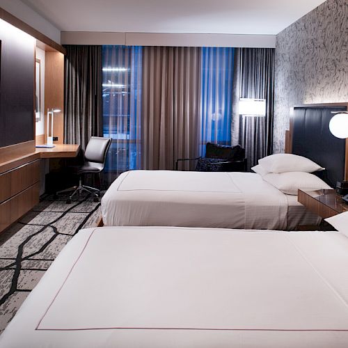 A modern hotel room with two double beds, a wall-mounted TV, a work desk and chair, soft lighting, and decorative features on the wall.