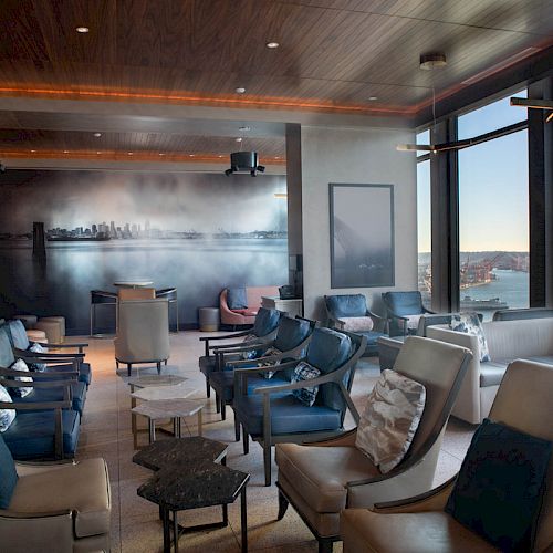 A modern lounge with numerous chairs, tables, and large windows providing a scenic waterfront view on a sunny day, featuring a cityscape mural.