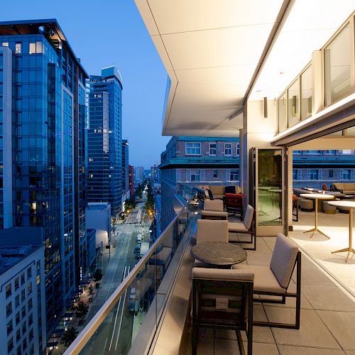 The image shows a modern city skyline at dusk with tall buildings and a spacious balcony featuring outdoor furniture and tables.