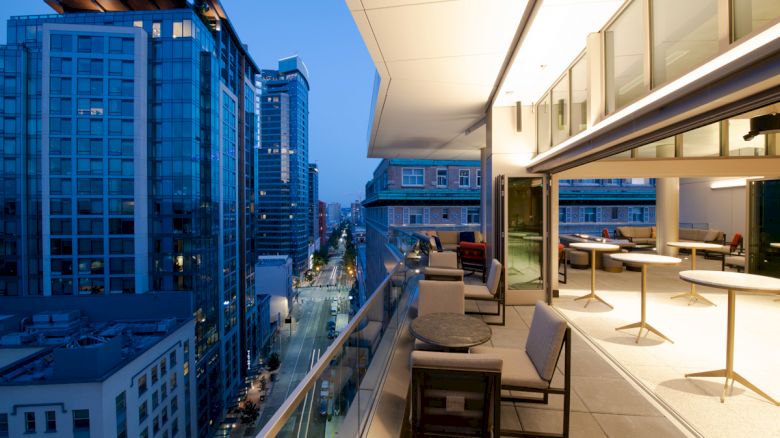 A modern outdoor balcony with seating and tables is overlooking a city street lined with high-rise buildings during the evening.