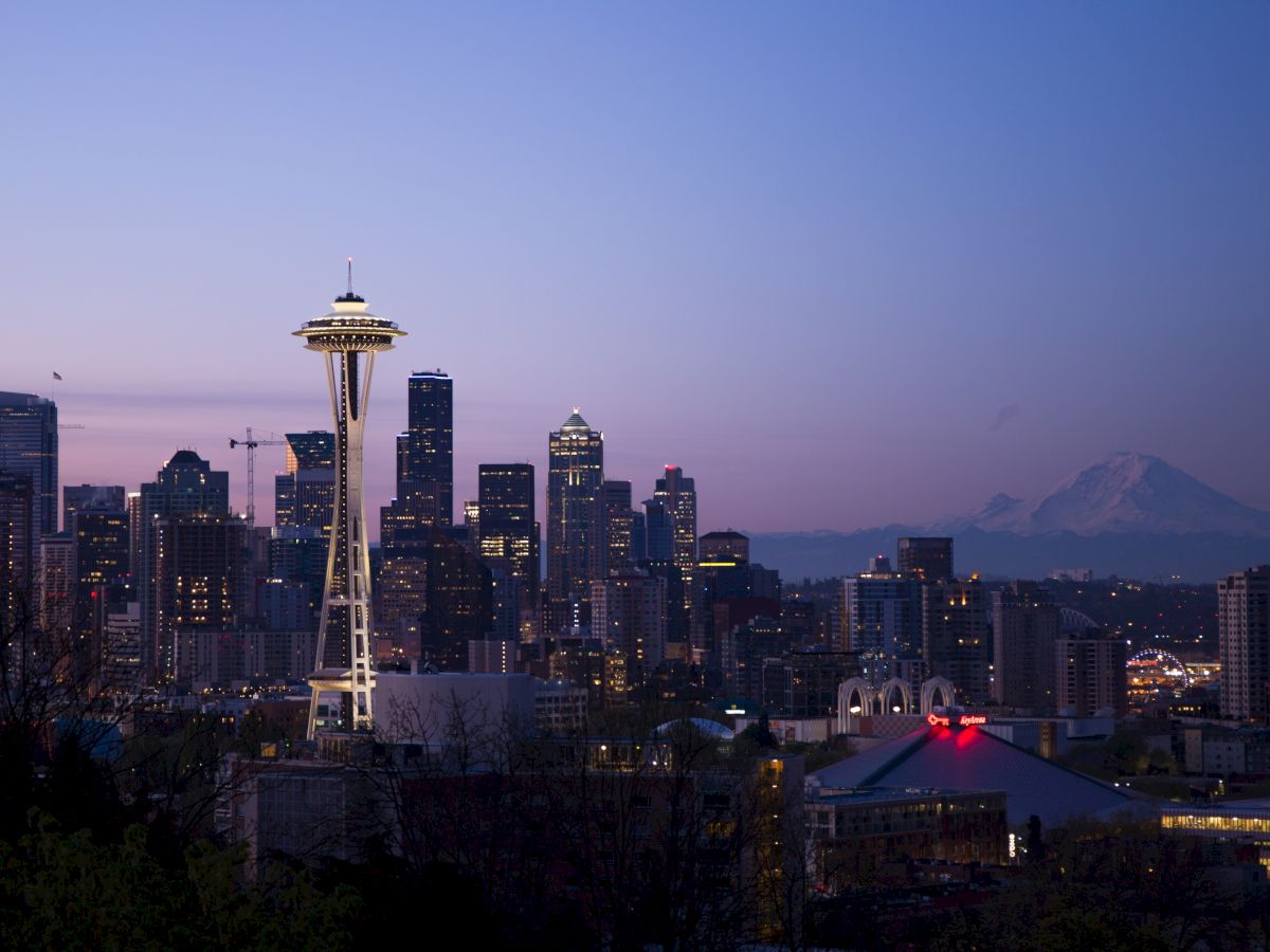 A cityscape at sunset features the Space Needle, various buildings, and Mount Rainier in the background, under a clear sky with fading daylight.