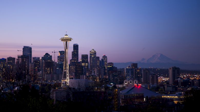 A cityscape at sunset features the Space Needle, various buildings, and Mount Rainier in the background, under a clear sky with fading daylight.