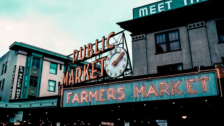 The image shows a neon sign for a Public Market and Farmers Market against a building backdrop, with a clock and partial hotel sign visible.