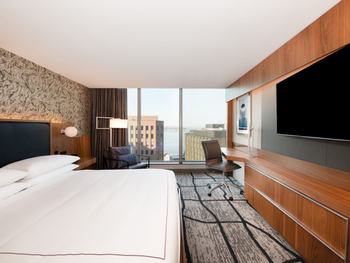 A modern hotel room with a large bed, desk, chair, and a flat-screen TV, featuring a view of city buildings through the window and stylish decor.