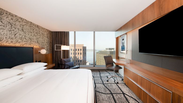 A modern hotel room with a large bed, desk, chair, and a flat-screen TV, featuring a view of city buildings through the window and stylish decor.
