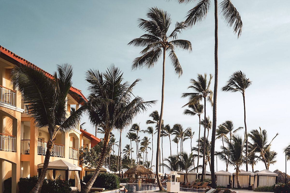 A serene poolside scene featuring lounge chairs, tall palm trees, and a multi-story building under a clear sky, evoking a relaxing tropical getaway.