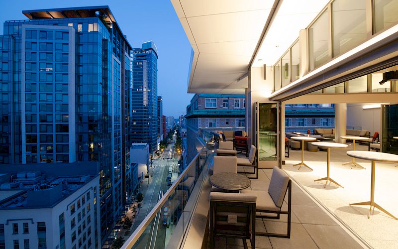 A modern cityscape during twilight with tall buildings, and an open terrace featuring tables and chairs for relaxed outdoor seating.