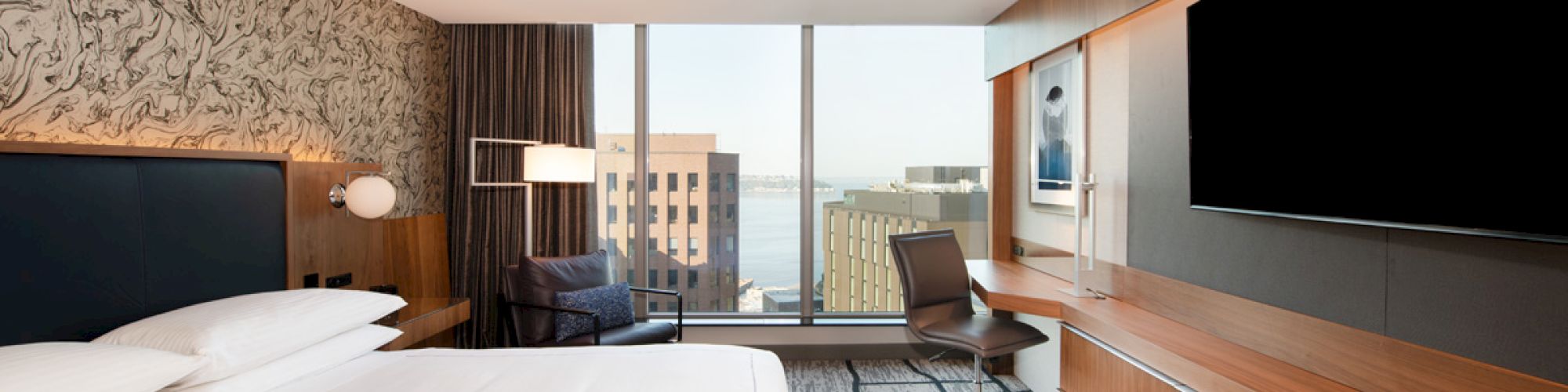 The image shows a modern hotel room featuring a large bed, a TV mounted on a wooden panel, a desk with a chair, and a window with a city view.